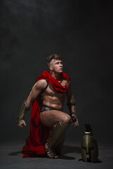 Fototapeta na wymiar A young, athletic man dressed as a Roman warrior in a red cloak and helmet on a black background dropped to one knee.