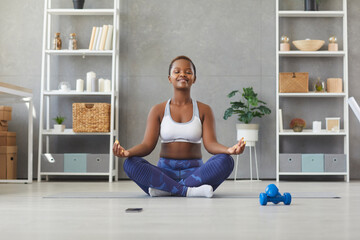 Happy calm young African American woman in sportswear sit in mudra pose asana meditate at home. Smiling millennial biracial girl practice yoga have meditation online class session. Sport concept.