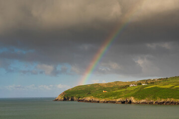 View on Mizen Peninsula in county Cork, Ireland. Colorful rainbow symbol of luck in the sky. Calm water of Atlantic ocean and green fields. Irish landscape. Popular travel destination. Nature wonder. - obrazy, fototapety, plakaty
