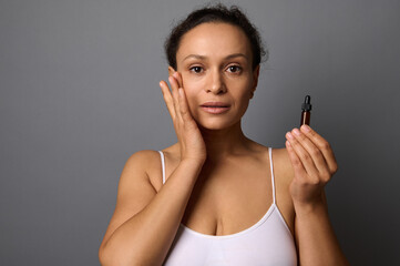 Beautiful middle aged woman holds a jar with beauty product, applies it on her face and performs anti-aging lymphatic drainage massage for rejuvenation and skin smoothing. SPA, cosmetology concept