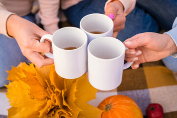 Women's hands with white mugs at a picnic. Friends drink hot tea at a picnic in autumn, close-up view from above. Selective focus