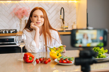 Medium shot portrait of smiling attractive redhead woman vlogger shooting video food blog about cooking on camera of mobile phone, sitting at kitchen with modern light interior, selective focus.