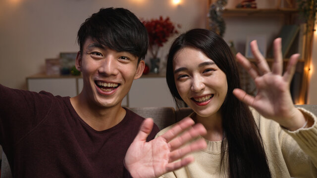 Front View Young Asian Man And Woman Sitting On Couch At Home And Face Camera With Video Call To Family. Cheerful Lovely Couple Celebrating Anniversary Indoors And Waving Hands To Lens And Chatting