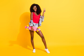 Full length body size photo of girl keeping skate board showing v-sign gesture isolated on vibrant yellow color background