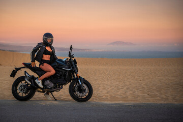 Fototapeta na wymiar Mujer joven sexy y atlética sentada en su moto al atardecer en las dunas de Tarifa, Cádiz, Andalucía, España