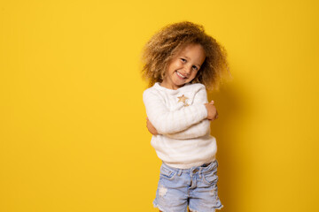 Portrait of charming little girl embracing herself and smiling isolated over yellow background.