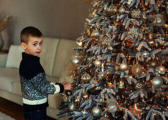 A boy decorating xmas tree in the evening at home. Serious work-focused child boy in a knitted sweater look at camera. Family with kids celebrate winter holidays. The eve of Christmas