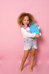 Beautiful little girl with curly hair holding skateboard isolated over pink background.