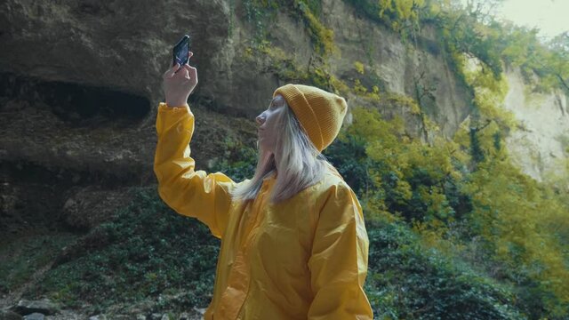Caucasian girl in a yellow raincoat and a hat takes pictures of nature on the phone. Travel to the mountains.