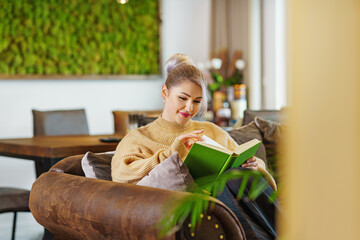Young beautiful woman relaxing in cozy green apartment, reading a book, wearing warm knitted...