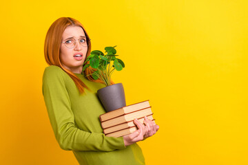 Profile photo of dislike ginger hair young lady hold books plant wear eyewear green sweater isolated on yellow color background