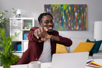 Elegant man sits at desk in corporate company, coffee mug laptop, smiling dark-skinned guy stretches hand out in front of him points at someone towards the camera, selects a person, happy