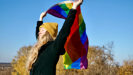 Portrait of a young, Caucasian, blonde, girl with acne holding a rainbow LGBT flag in autumn. Woman in yellow hat and black coat on a sunny day