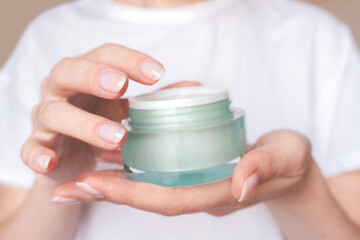 Female hands with natural pink manicure holding a transparent cream jar. Woman in white holds beauty product. Concept of body care and skin protection. Moisturizing lotion closeup. Facial skincare