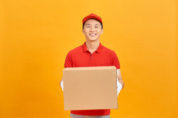 Image of a happy young delivery man in red cap standing with parcel post box isolated over yellow background.