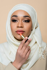 young muslim woman in hijab applying lipstick isolated on beige