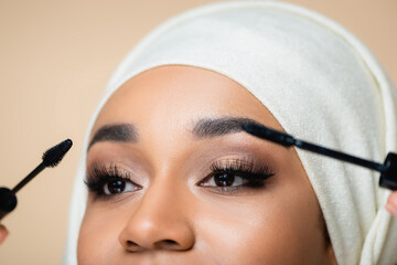cropped view of muslim woman in hijab applying mascara with brushes isolated on beige