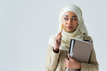 pensive muslim woman in hijab holding pencil and folders isolated on grey