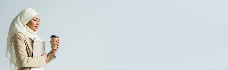 Muslim businesswoman in hijab holding folders and paper cup isolated on gray, banner