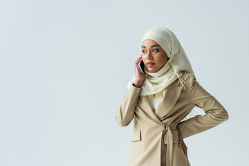 stylish muslim woman in hijab and beige suit talking on smartphone isolated on grey