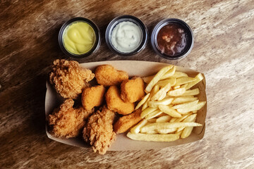 Top view of chicken nuggets, batter wings and fries. A set of three dishes in a paper box. Fast food cafes and restaurants. The food is laid out in a row. Three types of sauce
