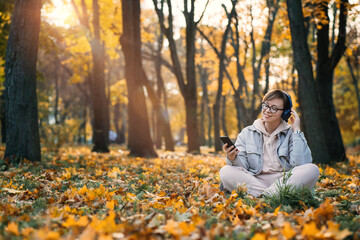 Caucasian middle aged woman in earphones listening to music, meditation app on smartphone and meditating in lotus pose at autumn park. Meditation app, Mental health, self care, mindfulness concept