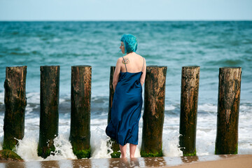 Young blue-haired woman in long dark blue dress standing on sandy beach looking at sea horizon
