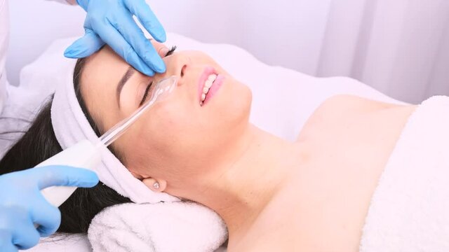 Professional beautician in medical gloves doing the rejuvenate procedures to a young woman, smoothes wrinkles using cosmetic instrument, electrical appliance.