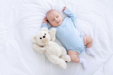 baby boy sleeps on the bed lying on his back with a soft toy bear in blue pajamas holding his hands...