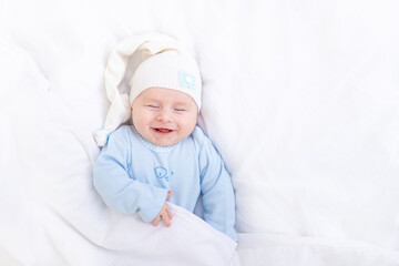 baby boy smiling in his sleep on the bed under the blanket in a cap, healthy newborn sleep