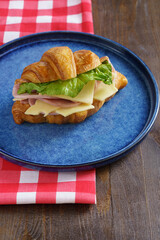Croissant with ham, cheese, cucumber and herbs on a wooden kitchen table on a blue plate and a checkered red tablecloth. . High quality photo