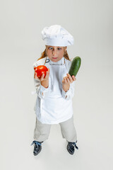 High angle view of little cute girl in white cook uniform and huge chef's hat posing isolated on white studio background.