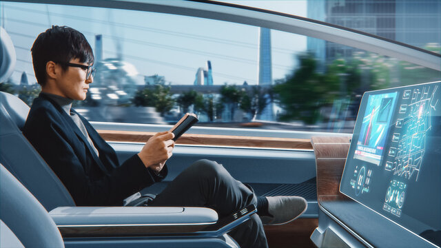 Futuristic Concept: Handsome Stylish Japanese Businessman in Glasses Reading Notebook and Watching News on Augmented Reality Screen while Sitting in a Autonomous Self-Driving Zero-Emissions Car. 