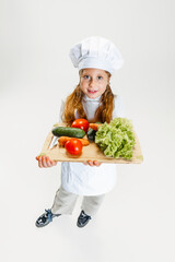 High angle view of little cute girl in white cook uniform and huge chef's hat holding tray with healthy vegetables isolated on white studio background.