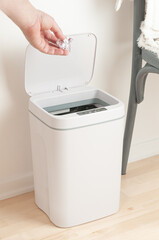 Smart recycle bin. Electronic gadget for home and office. A man's hand throws out trash. Stands on a wooden floor. The background is white.