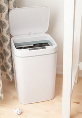 Smart recycle bin. Electronic gadget for home and office. Cover is open. Stands on a wooden floor. Next to the chair. The background is white.