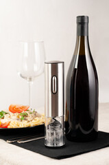 Electric corkscrew in gray metal. There is a bottle of wine, a plate of pasta, a glass and cutlery...