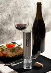 Electric corkscrew in gray metal. There is a bottle of wine, a plate of pasta, a glass and cutlery...