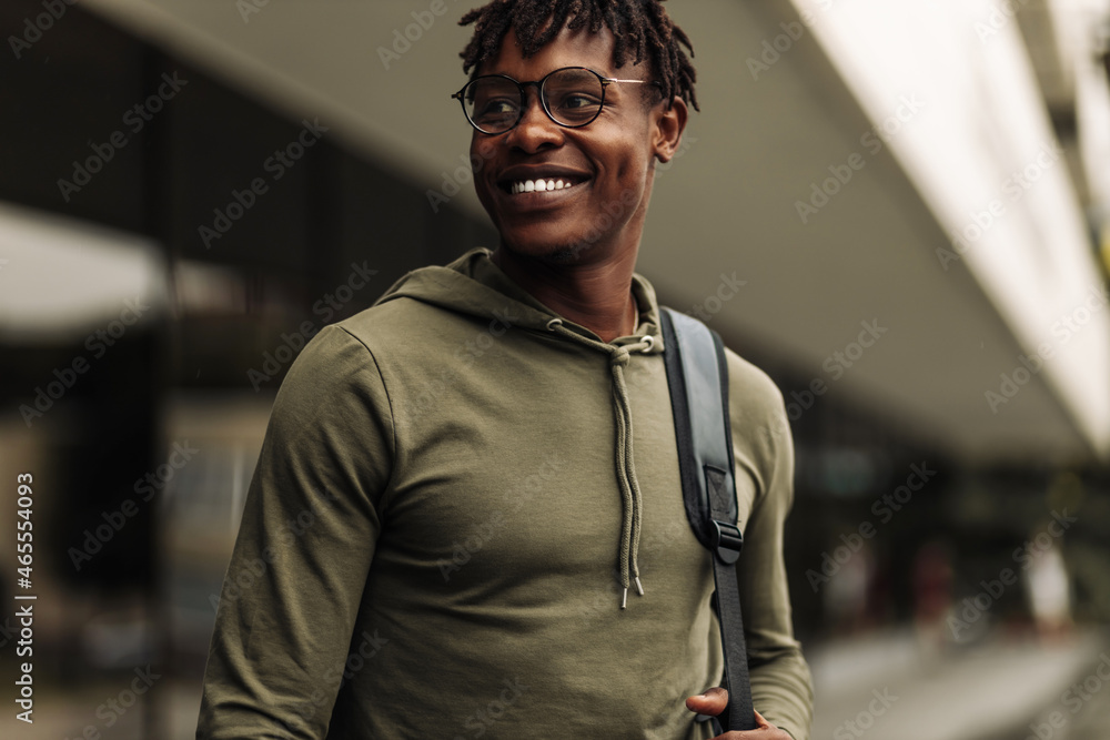 Wall mural Happy successful african student, wearing glasses and with a black backpack, standing on the steps and smiling