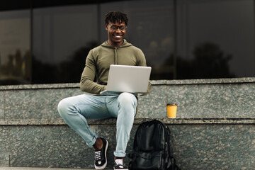 African man, using laptop sitting on city stairs, positive student studying online, outdoors