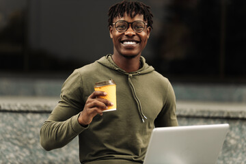 Young African American businessman working online with laptop sitting on bench holding takeaway coffee, outdoors