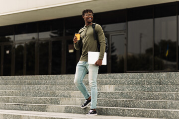 African American man, drinking coffee in a cup and walking up the stairs in the city center with a laptop in his hands