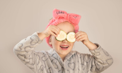 A little girl wearing bathrobe and holding pieces of cucumber on her eyes