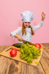 Young little girl in white cook uniform and huge chef's hat cutting fresh vegetables isolated on pink studio background.