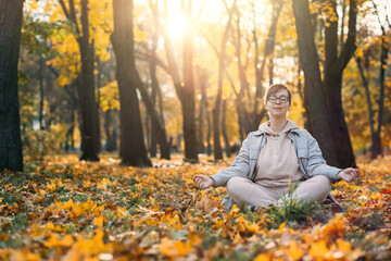 Caucasian middle aged woman meditating in lotus pose at autumn park with sunlight. Yoga at fall park. Woman in glasses relaxing on yellow leaves. Meditation, Mental health, self care, mindfulness