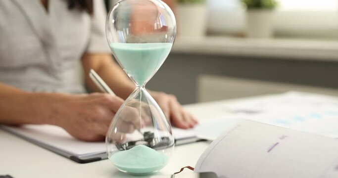 Woman fills out paperwork against background of hourglass at workplace 4k movie