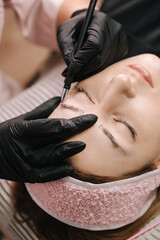 Close-up girl's face with closed eyes. The hand of a master beautician draws a sketch of an eyebrow with a black pen on the girl's skin before microblading