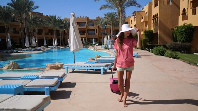 Beautiful woman in big white hat and pink travel bag walking on territory of hotel near swimming pool. She walks on alley with palm trees. HD video in ProRes 1920x1080