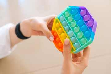 Close-up top view of of unrecognizable young woman playing with rainbow pop-it fidget toy sitting at table. Closeup of female pushing colorful iridescent soft silicone bubbles sitting at desk.