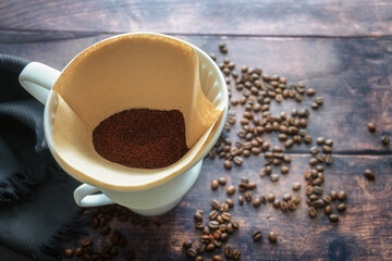 Ground coffee in a paper filter bag and in a porcelain holder on a mug for an aromatic drip brewed hot drink, rustic wooden table with some beans, copy space, selected focus